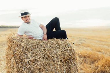 Şapkalı yakışıklı genç adam saman balyasında yatıyor ve tarlada kameraya gülümsüyor.