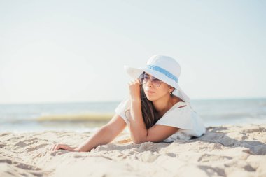 Güneşli yaz gününde kumlu sahilde uzanan şapkalı ve güneş gözlüklü çekici genç bir kadın.