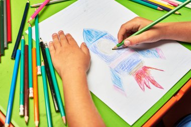 Küçük çocuk masada kalemle roket çiziyor.