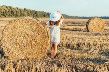 Tarlada saman balyasının yanında duran beyaz elbiseli ve şapkalı çekici genç bir kadın.