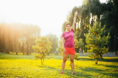 Spor giyimli genç bir kadın sabah parkta yeşil çimenler üzerinde egzersiz yapıyor.