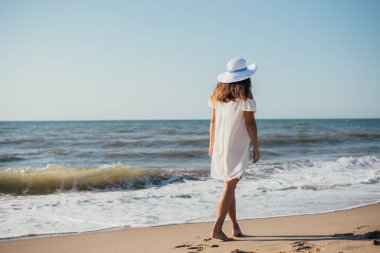 Şapkalı genç bir kadının kumsalda yürüyüşünün arka görüntüsü.