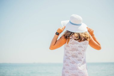 Beyaz elbiseli ve şapkalı genç bir kadının kumsalda durup denize bakışı.