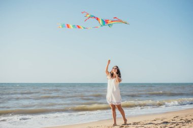 Çekici, neşeli, genç bir kadın renkli bir uçurtma tutuyor ve kumsalda yürüyor.