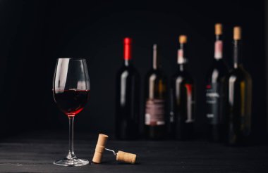 yakın plan cam, kırmızı şarap, tirbuşon ve siyah arka planda şarap şişeleri.