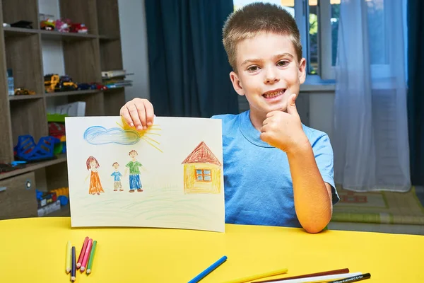 小男孩在桌旁用铅笔画家庭和房子 — 图库照片