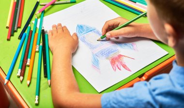 Küçük çocuk masada kalemle roket çiziyor.