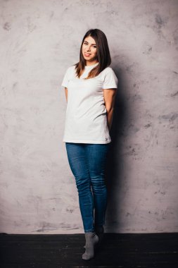 Gri duvarın yanında durup kameraya gülümseyen beyaz tişörtlü ve kot pantolonlu güzel bir kadın.
