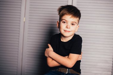 Siyah tişörtlü ve kot pantolonlu sevimli küçük çocuk taburede oturup kameraya bakıyor.