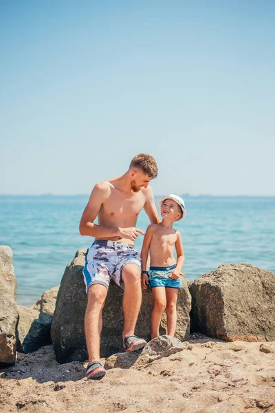 快乐的父亲和儿子穿着泳衣坐在沙滩上的岩石上 — 图库照片