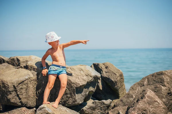 Chłopiec Szortach Kapeluszu Siedzi Skale Wskazuje Palcem Wybrzeżu Morza — Zdjęcie stockowe