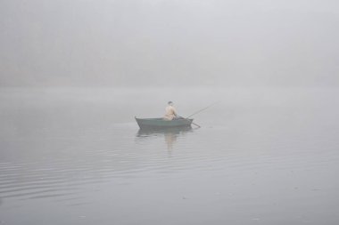 silhouette of fisherman in boat floating on calm lake at foggy morning