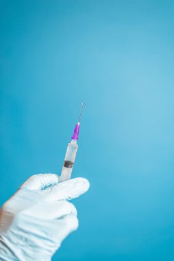Eldivenli doktorun mavi arka planda aşısı olan bir şırınga tutuşu. Enjeksiyon solüsyonlu şırınga. Sıvı ilaç veya uyuşturucu