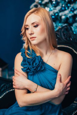 Mavi elbiseli çekici sarışın kadın Noel ağacının yanında oturuyor.