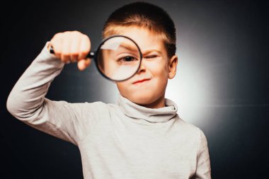 Küçük sevimli çocuk büyüteç tutuyor ve gri arka plandaki kameraya bakıyor. 