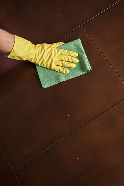 cropped shot of woman in rubber glove holding rag and cleaning brown surface at home