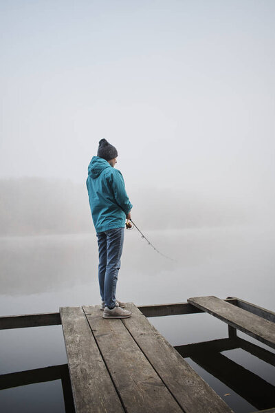 back view of young woman standing on wooden pier and fishing with rod at foggy morning