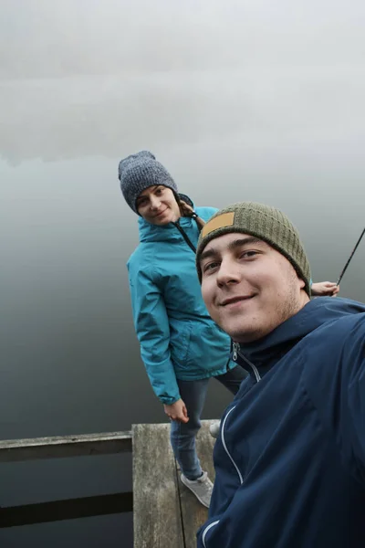 朝霧の中で川の近くで一緒に釣りをしながらカメラで微笑む幸せなカップル — ストック写真
