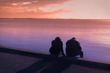 Gün batımında deniz kenarında oturan bir çiftin silueti.