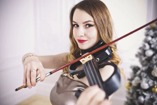 Uma menina bonita com lábios vermelhos toca um violino elétrico. Close-up. Estúdio Retrato com fundo de árvore de natal — Fotografia de Stock