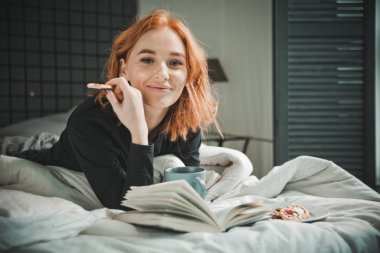 Modern apartmandaki beyaz yatakta mutlu sabahlar inanılmaz neşeli kız. Gri pijamalı, uzun bacaklı, kızıl saçlı, okuma kitabı olan hoş bir genç kadın. Neşeli zaman, heyecanlı duygu