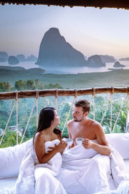 couple waking up in bed in nature jungle looking out over ocean during sunrise at wooden hut in the moutains of Thailand