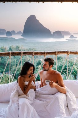 couple waking up in bed in nature jungle looking out over ocean during sunrise at wooden hut in the moutains of Thailand