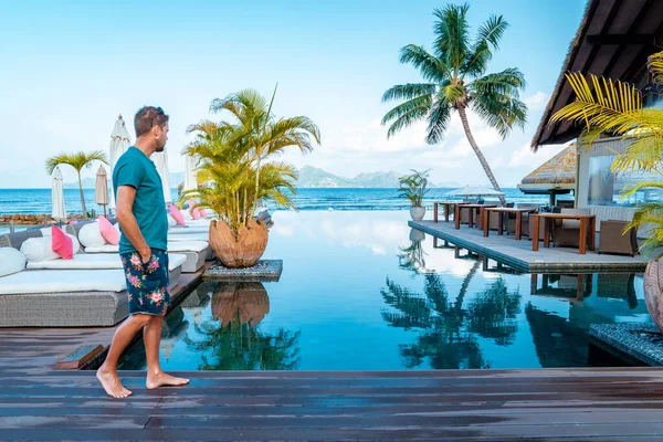 Luxury swimming pool in tropical resort, relaxing holidays in Seychelles islands. La Digue, Young man during sunset by swimpool — Zdjęcie stockowe