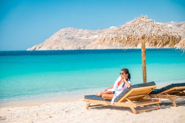 Yaz boyunca şemsiyeli ve lüks plaj koltuklu Mykonos plajı, Elia sahilinde dağlı mavi okyanus Mikonos Yunanistan