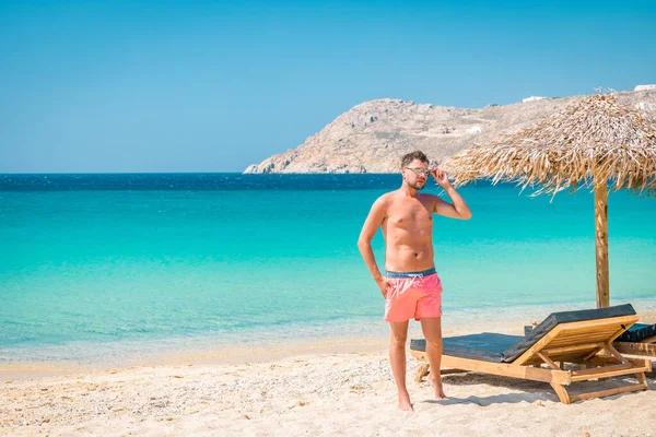 Tatilde yüzen adam Mykonos, yaz boyunca şemsiyeli ve lüks plaj koltuklu Mykonos plajı, Elia sahilinde dağlı mavi okyanus Mikonos Yunanistan — Stok fotoğraf