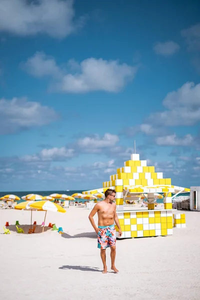 Miami Beach Florida, hombres jóvenes en la playa con cabaña de salvavidas colorido, hombre caminando en la playa en Miami durante las vacaciones — Foto de Stock