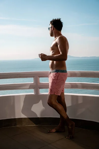 guy drinking coffee at the balcony with sea view, men in swim short drinking coffe ocean view on balcony