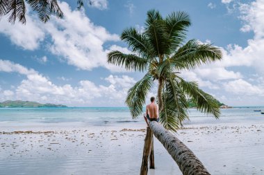 Cote DOr Sahili, Praslin Seyşeller tropikal adası, beyaz sahilde yüzen genç erkekler büyük kayalar ve kayalar ve tatil sırasında palmiye ağaçları olan beyaz tropikal bir plaj.