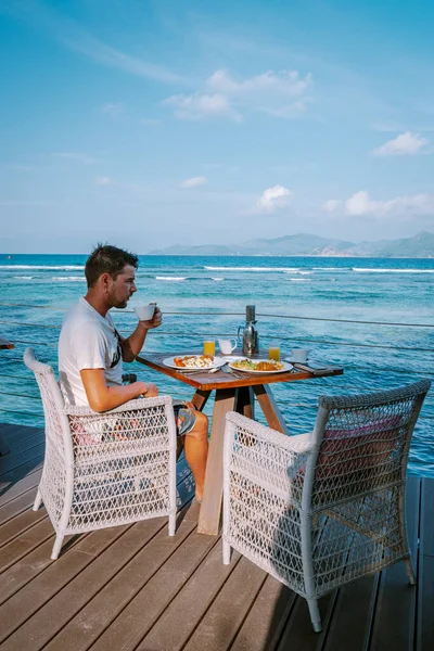 Genç adam, tatil sırasında okyanusa bakarak kahvaltı yapıyor. Seyşeller LA Digue Adası. — Stok fotoğraf