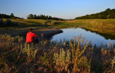 Kırmızı tişörtlü yalnız bir adam kuruyan bir nehrin kıyısında oturuyor, küresel ısınma, iklim değişiklikleri