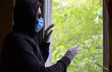 Koyu renk kapüşonlu, güneş gözlüklü, mavi cerrahi antiviral maskeli ve cama dokunan ve pencereden yeşil ağaca bakan yalnız üzgün adam.