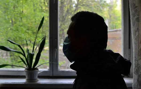 silhouette of a lonely sad man in surgical antiviral mask on self-isolation sitting in a room in front of a window