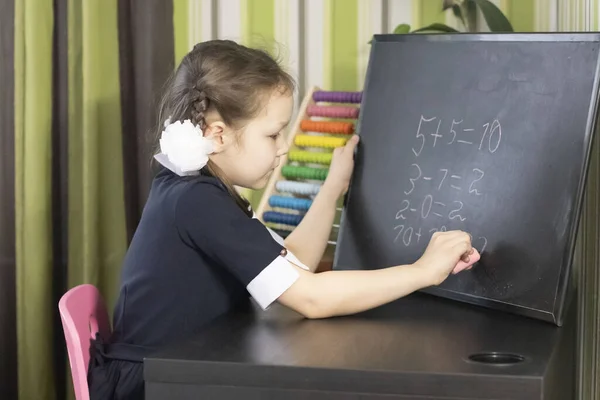 Distance learning online education. Girl writes math in chalk on a blackboard. Schoolgirl studying at home at the table. Examples from elementary school, addition and subtraction.