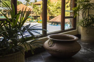 Yeşil bitki ve havuz manzaralı pencerenin yanında büyük bir bakır kap.