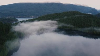 Crystal clear lake with small cloud-like fog, green forest around and mountains on the background