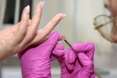 Pembe eldivenli manikürcü çeşitli ekipmanlarla yetişkin bir müşterinin tırnaklarını inceliyor. Manikür. Güzellik salonu. Eldivenli eller ellerin tırnaklarıyla ilgilenir. Prosedür.