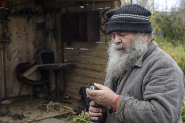 An elderly gray bearded man in a headdress and a worn jacket is engaged in construction work in his yard