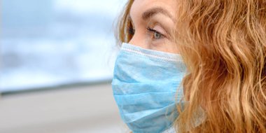 Tıbbi maske takan kızıl saçlı yetişkin bir kadının yüzü. COVID-19, grip, SARS, alerji, virüslerden korunma. Şehirdeki hava kirliliği kavramı. Kapat..