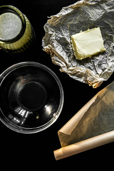 Ingredientes y utensilios para hacer galletas y cocinar. Mantequilla amarilla, azúcar en un plato de cristal sobre una mesa negra. La vista desde arriba —  Fotos de Stock