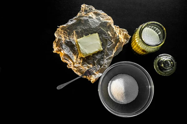 Ingredientes e utensílios para fazer biscoitos e cozinhar. Manteiga amarela, açúcar em um prato de vidro em uma mesa preta. A vista de cima — Fotografia de Stock