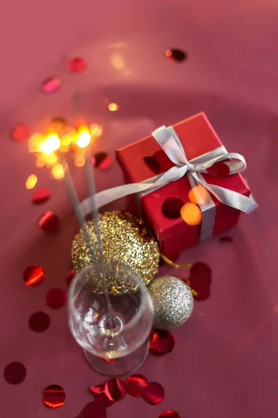 Christmas and new year sparklers in champagne glass on a red, pink, maroon background. New year and Christmas cards, colorful silver and gold baubles with a gift box and confetti. selective focus. — Stock Photo, Image