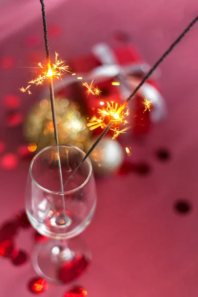 Christmas and new year sparklers in champagne glass on a red, pink, maroon background. New year and Christmas cards, colorful silver and gold baubles with a gift box and confetti. selective focus.
