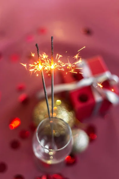 Christmas and new year sparklers in champagne glass on a red, pink, maroon background. New year and Christmas cards, colorful silver and gold baubles with a gift box and confetti. selective focus.