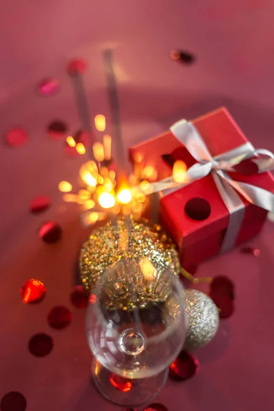 Christmas and new year sparklers in champagne glass on a red, pink, maroon background. New year and Christmas cards, colorful silver and gold baubles with a gift box and confetti. selective focus.