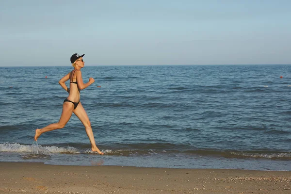 Mujer deportiva activa correr a lo largo del océano surf junto a la piscina de agua para mantenerse en forma y la salud. Atardecer arena negra playa de fondo con sol. Mujer fitness, ejercicio de jogging y actividad deportiva en vacaciones familiares de verano . — Foto de Stock